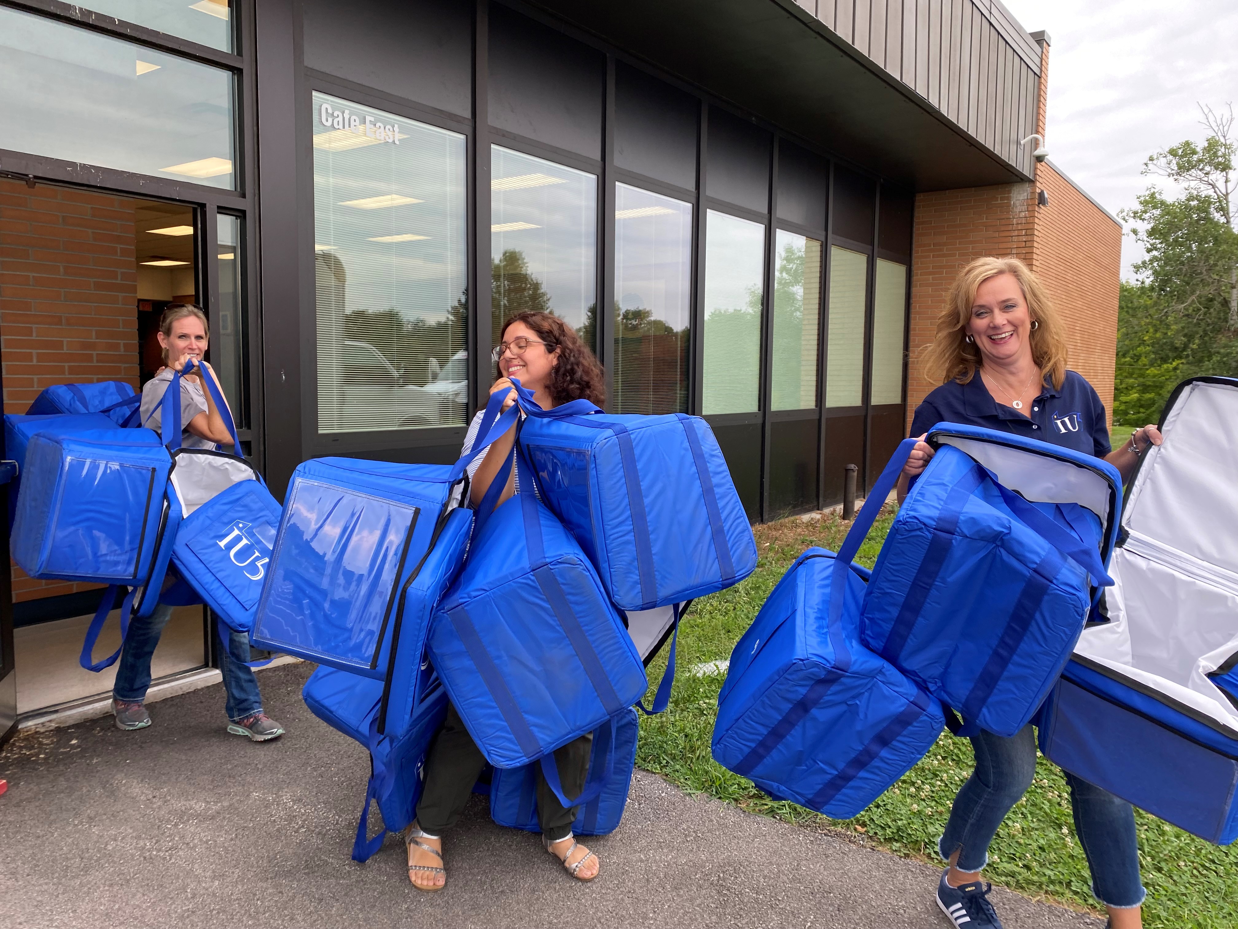 Three staff members walking out of a school. each holding multiple large empty cooler bags.