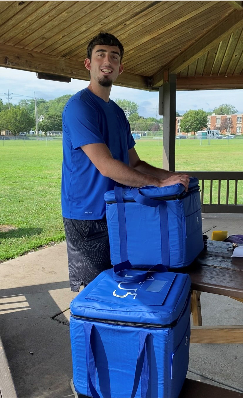 Paid volunteer, male college student, standing by coolers under pavilion.