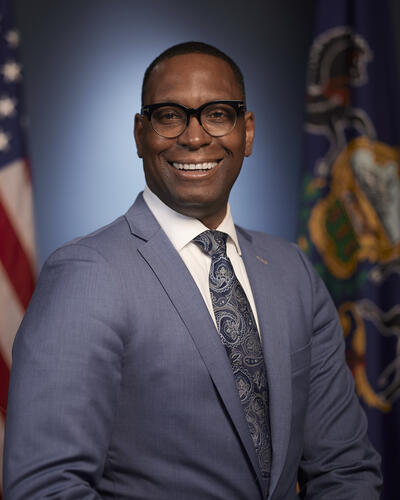 A portrait of Dr. Khalid Mumin  in a suit and tie smiling in front of an American and Pennsylvania state flag.